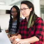 Two women working on a computer project together in an office setting.