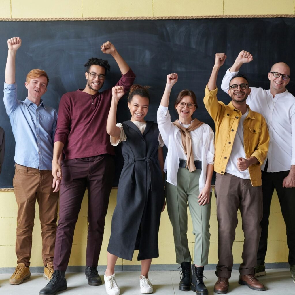 A group of six adults cheerfully standing with raised fists in front of a blackboard indoors.